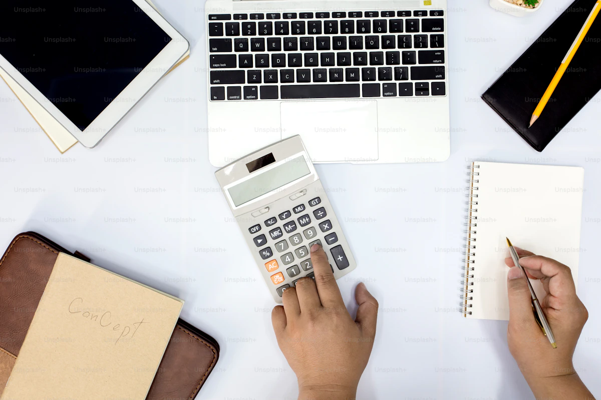 Person at a desk using a calculator and writing notes while monitoring financial information, representing the real cost breakdown of Trainerize